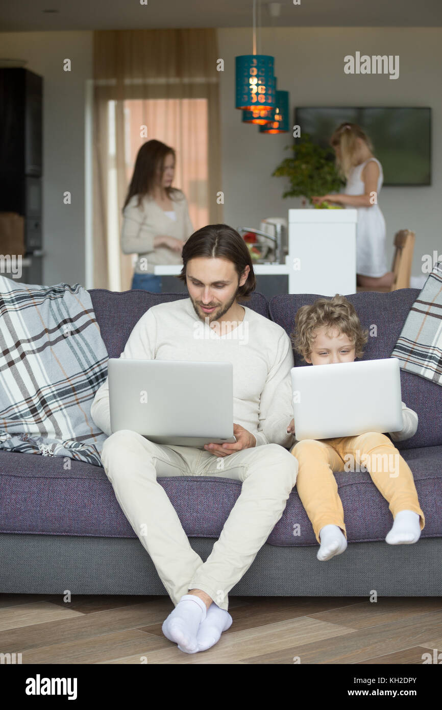 Dad and boy using laptops on sofa, kid imitating copying daddy holding computer sitting on couch while mother with daughter cooking spending time toge Stock Photo