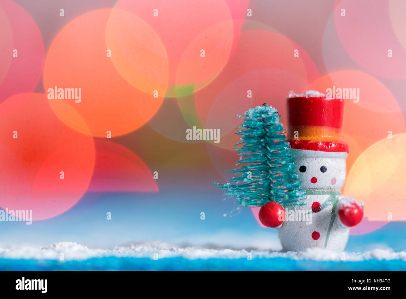 Snowman christmas decoration holding a small pine tree, snow on a blue wood surface, with background colorful bokeh lights, blue and cold tint Stock Photo