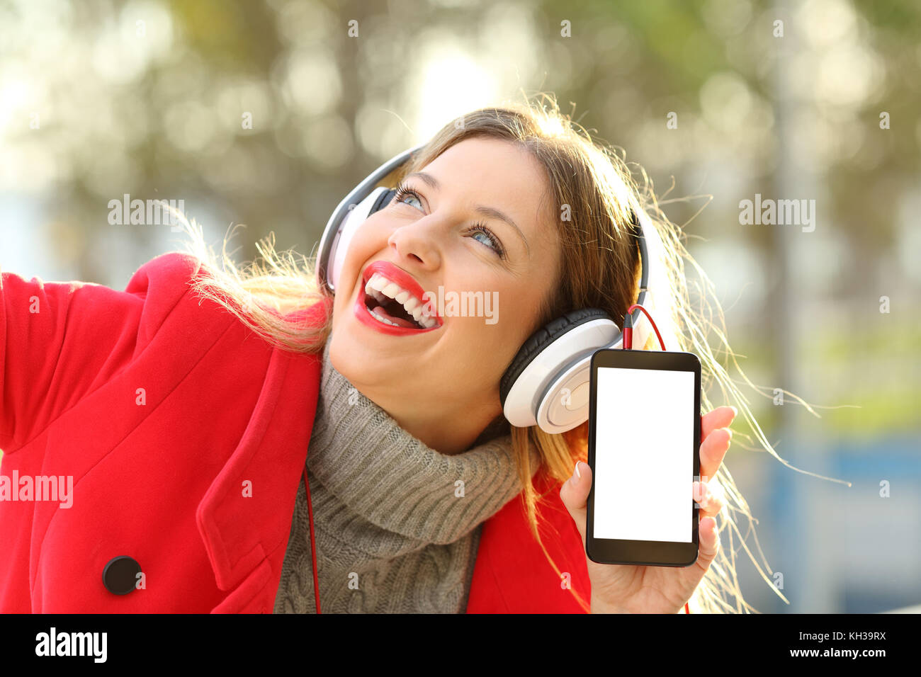 Happy girl wearing red jacket and headphones listening to music and showing smartphone screen in a park in winter Stock Photo