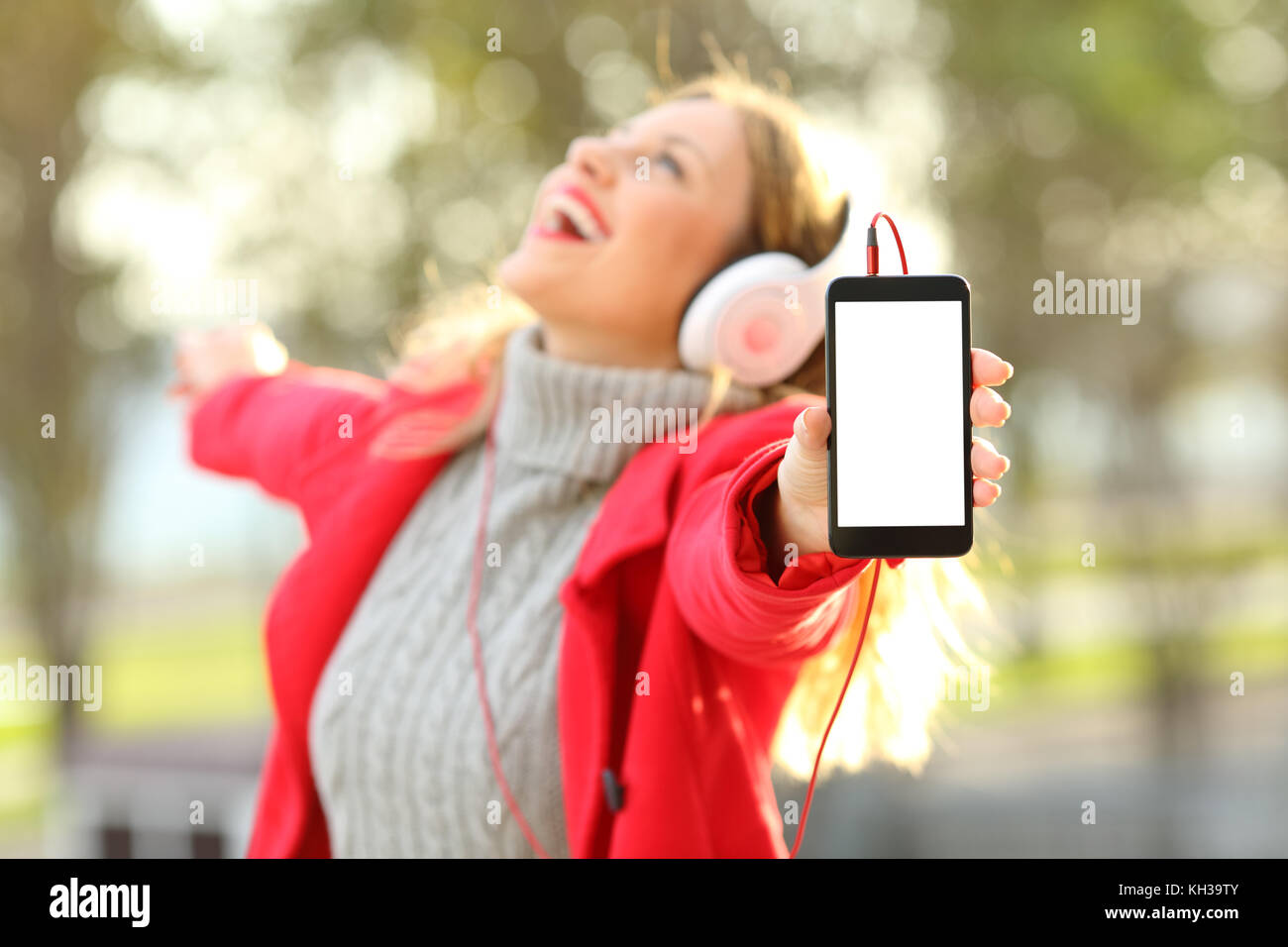 Joyful girl listening music and dancing with headphones showing a blank phone screen wearing a red jacket in winter Stock Photo