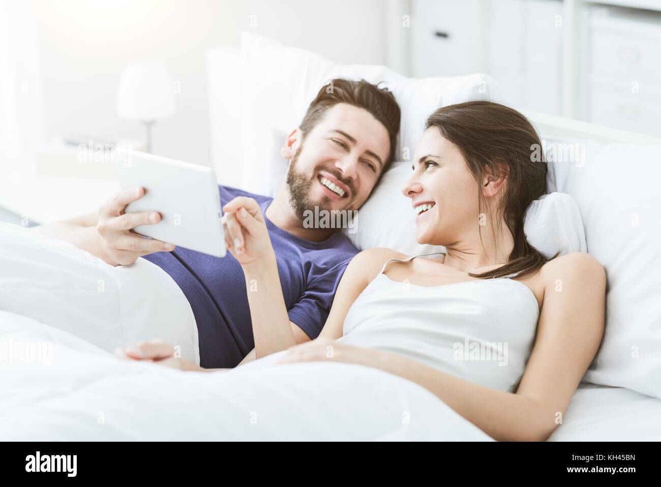 Young happy couple relaxing in bed and connecting with a touch screen ...