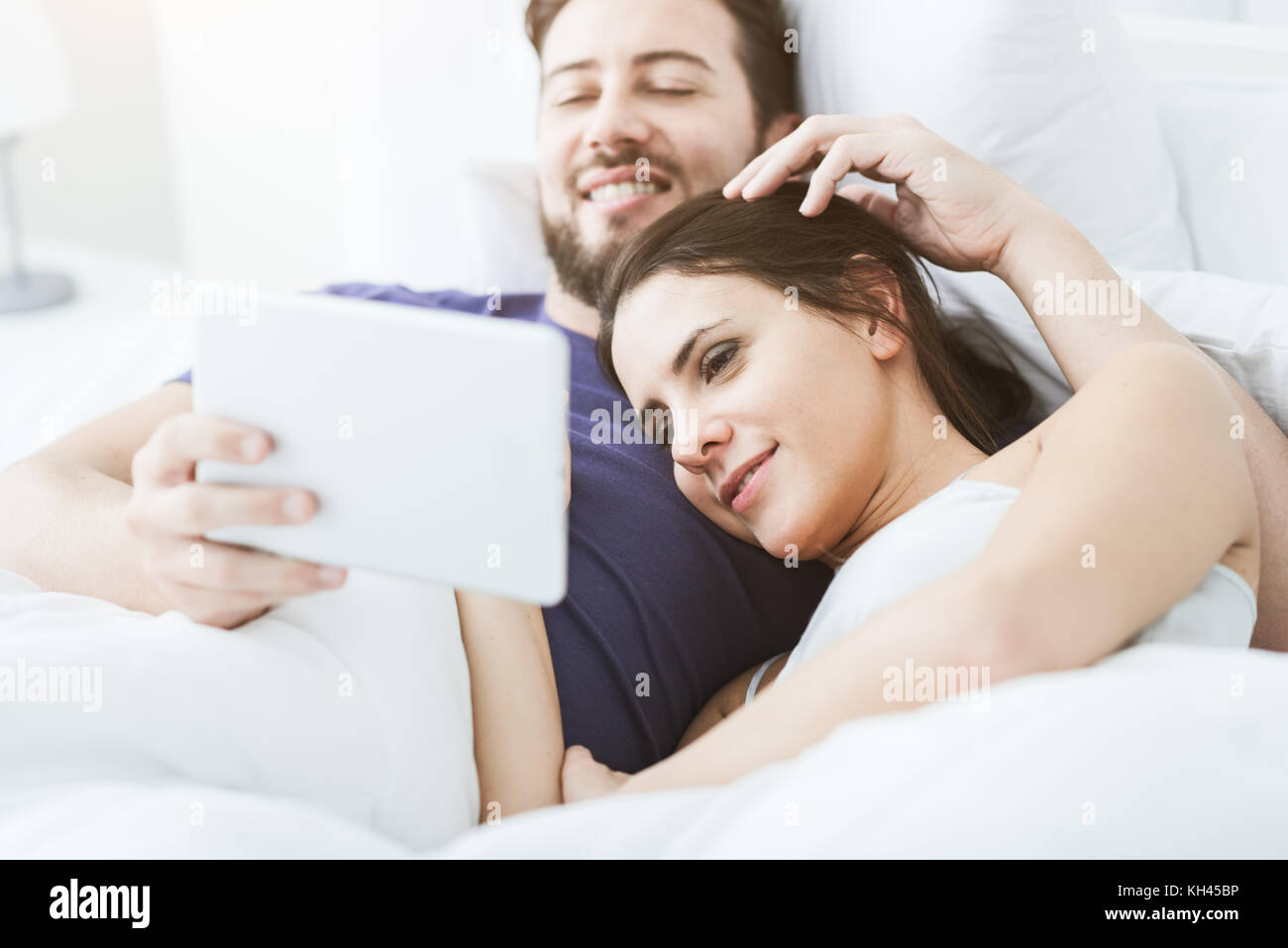 Young happy couple relaxing in bed and connecting with a touch screen ...