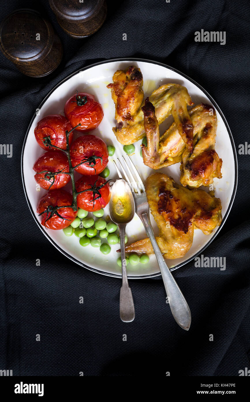Chicken wings, roasted cherry tomato and green peas. Healthy balanced meal on white plate over dark background. Top view dinner food Stock Photo