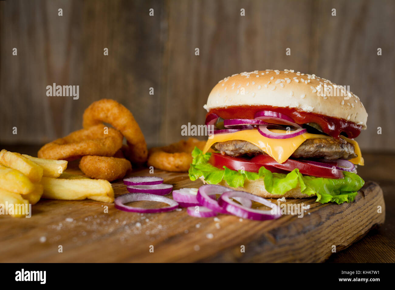 Cheeseburger, french fries and onion rings on wooden chopping board over wooden backdrop. Horizontal, closeup view Stock Photo