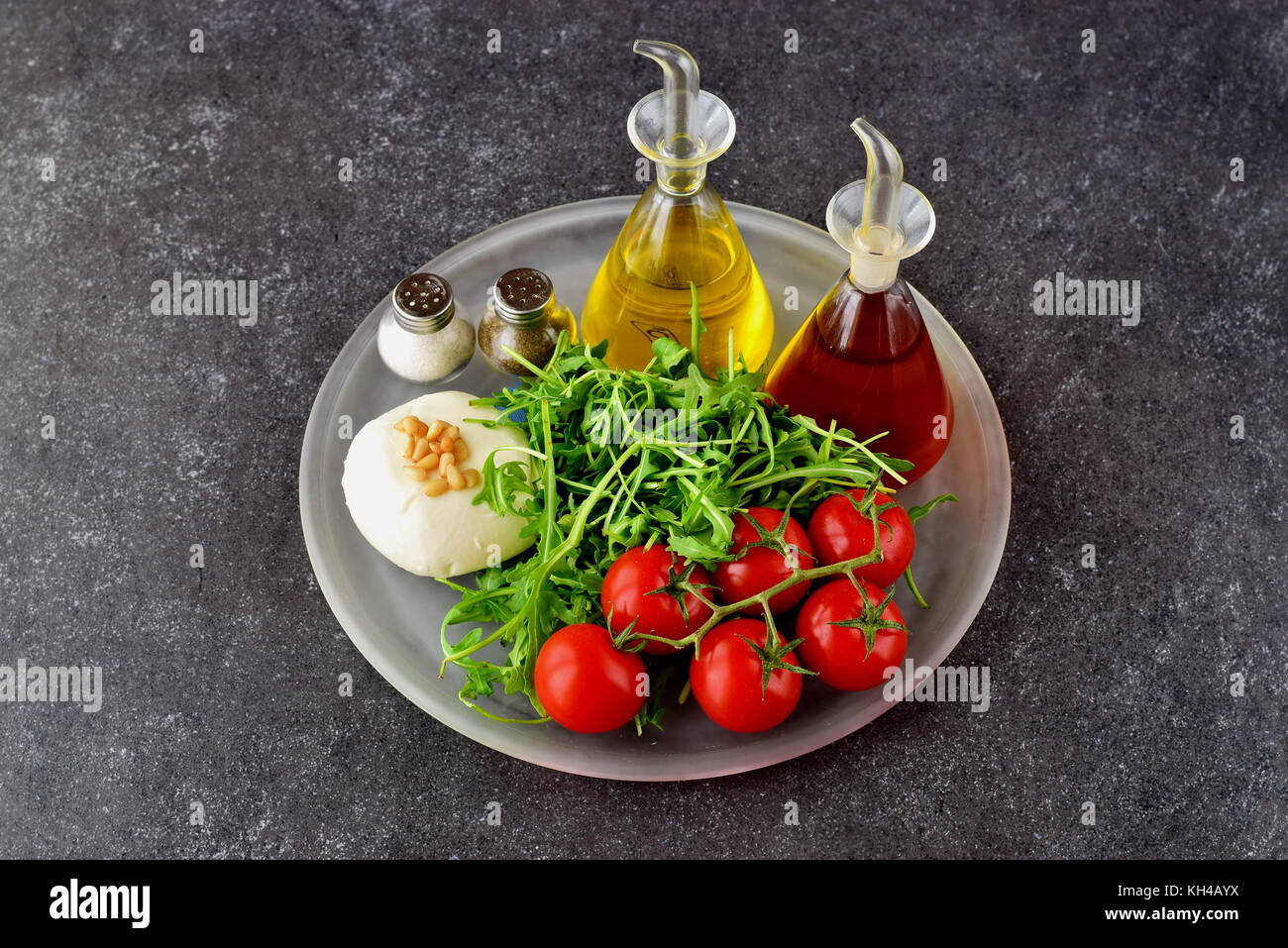 Ingredients for the traditional Italian caprese salad on a glass trey on a grey background with bottles of olive oil and wine vinegar. Stock Photo