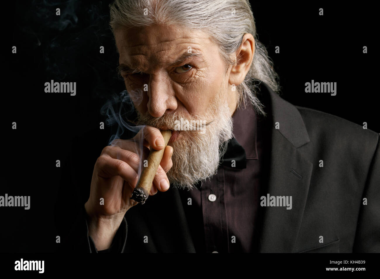 Stylish old man smoking cigar. Aged male in black suit in studio. Stock Photo