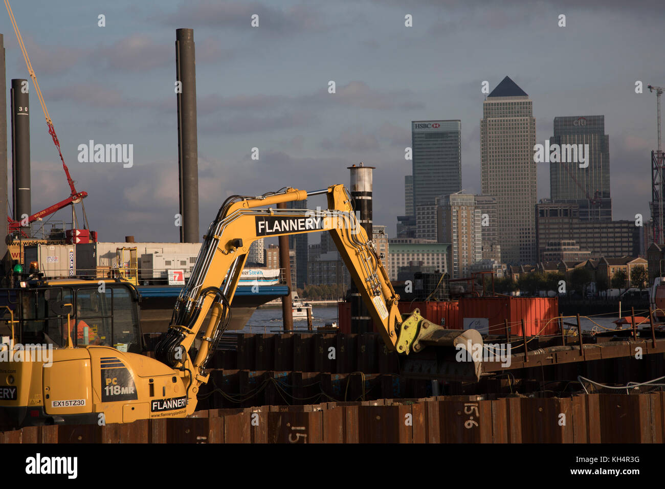 Construction work underway on the Thames Tideway Tunnel or Super Sewer on the River Thames near Wapping, with JCB diggers working in the foreground with Canary Wharf and the Docklands Financial District as the background in London, England, United Kingdom. The Thames Tideway Tunnel is an under-construction civil engineering project 25 km tunnel running mostly under the tidal section of the River Thames through central London, which will provide capture, storage and conveyance of almost all the combined raw sewage and rainwater discharges that currently overflow into the river. Stock Photo