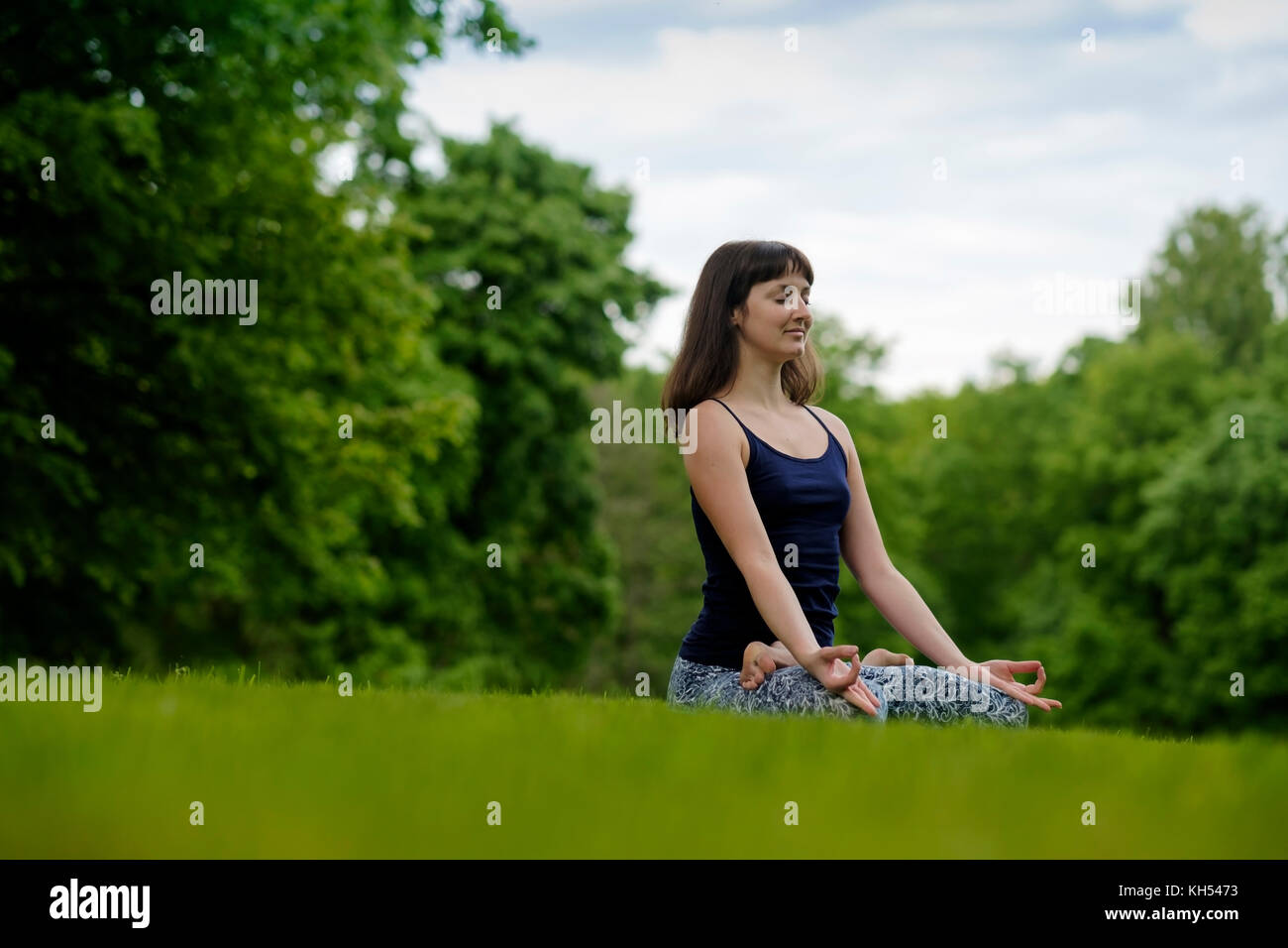 Beautiful young fit woman meditating, breathing, sitting with crossed legs in Lotus Posture in the park on summer day. Stock Photo