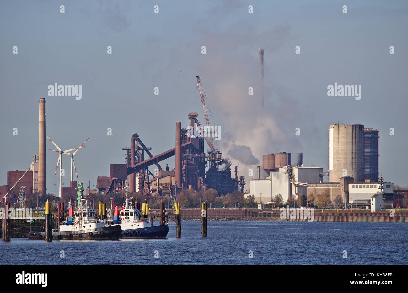 Two tug boats at their moorings with huge steel plant emitting clouds of grey smoke and wind power stations in the background Stock Photo