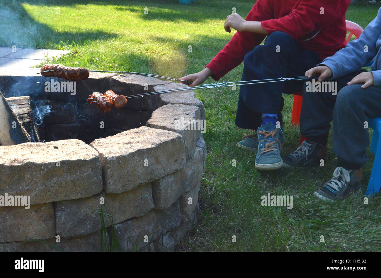 Two Boys Are Grilling Or Barbecuing Polish Sausages Over An Open