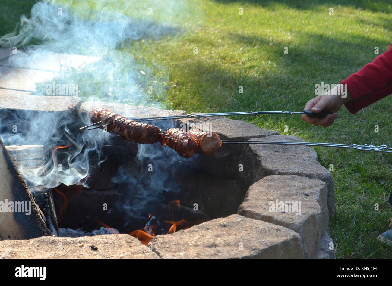 Two Boys Are Grilling Or Barbecuing Polish Sausages Over An Open