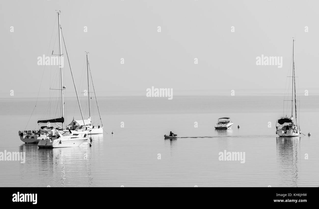 Man in dinghy driving from moored sailboats to shore. Sailboats, boats and ships are moored in calm bay in the morning. Stock Photo