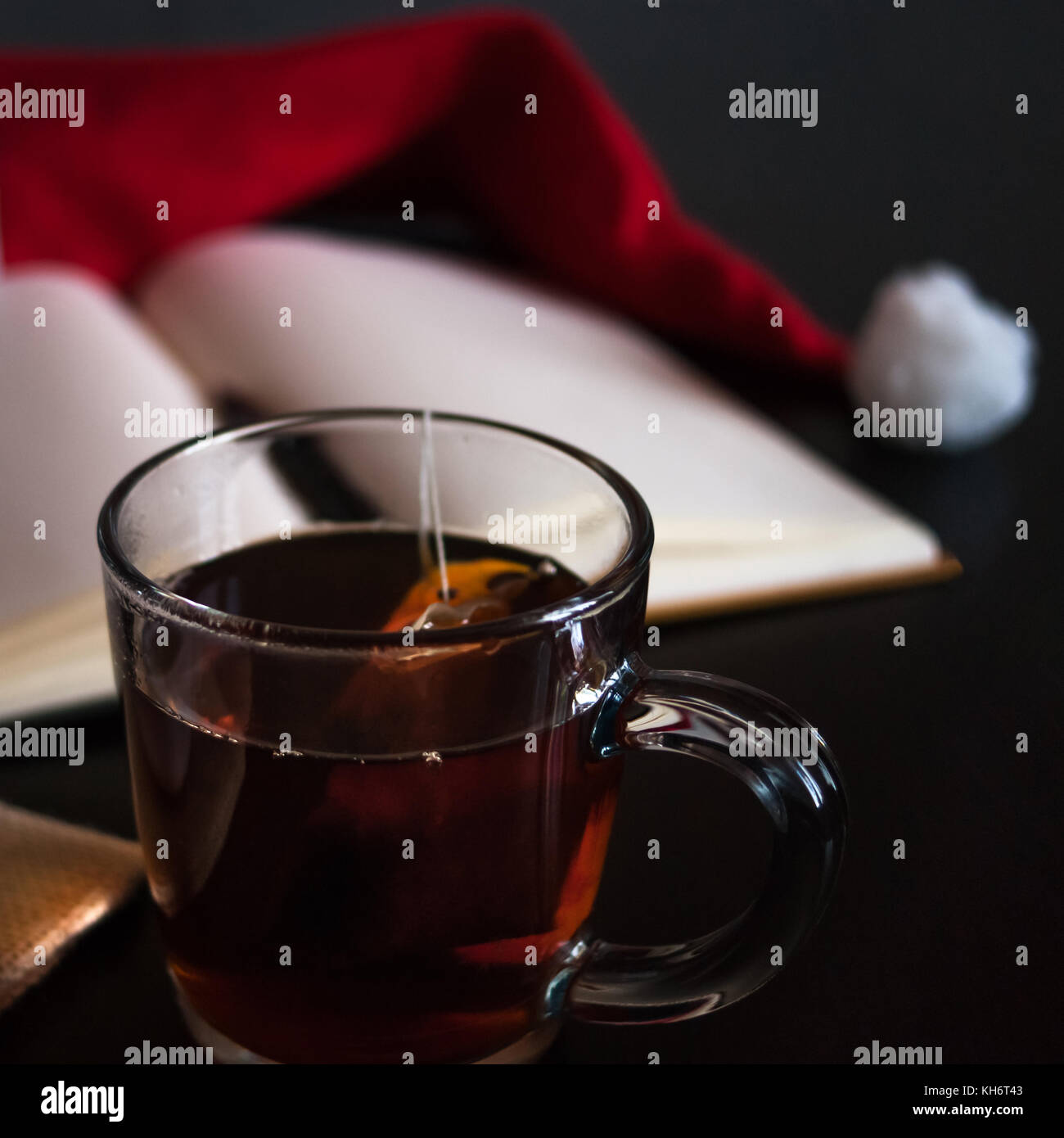 Planning future on Christmas concept: a notebook with blank pages, a black pen, Santa hat, glass mug with teabag, all on dark dining table Stock Photo