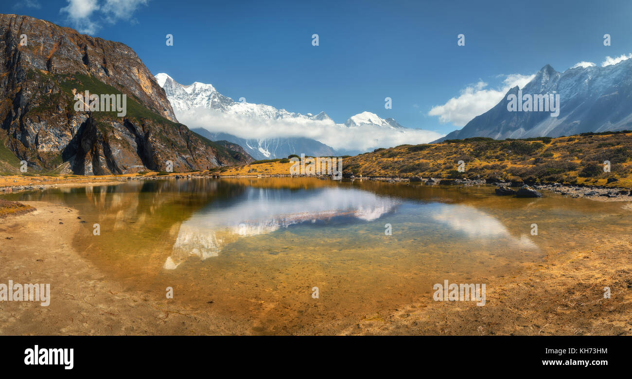 Beautiful view with high rocks with snow covered peaks, mountain lake, reflection in water, blue sky with clouds in sunrise. Nepal. Amazing panoramic  Stock Photo