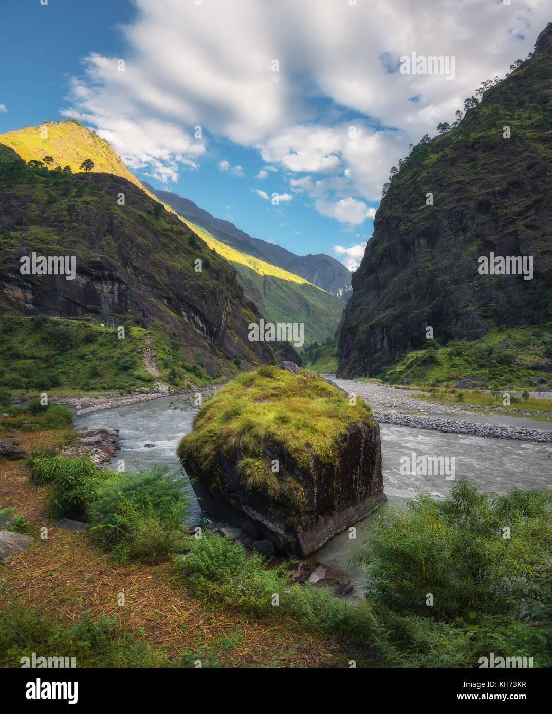 Amazing view with high Himalayan mountains, beautiful river, green forest, blue sky with clouds and big stone in water in autumn in Nepal at sunrise.  Stock Photo