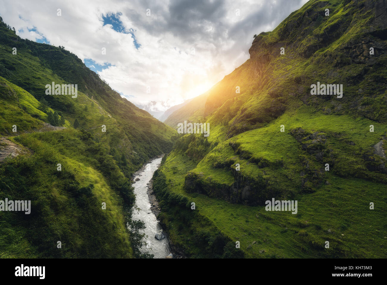 View with amazing Himalayan mountains covered green grass, river, meadows and forest, blue sky with clouds, sun and stones in water in autumn in Nepal Stock Photo