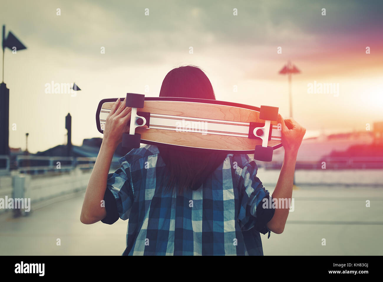 Young hipster woman holding skateboard behind head in sunset, outdoors, cinematic style, color graded Stock Photo