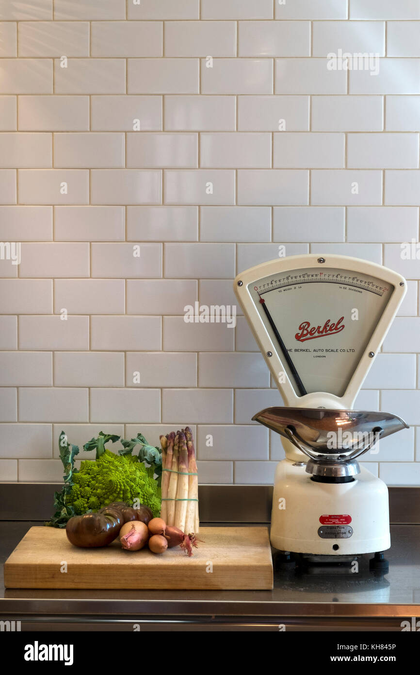 A few choice vegetables wait on a chopping board on a stainless steel kitchen work surface alongside a set of classic vintage grocers weighing scales made by The Berkel Auto Scale Co Ltd Stock Photo