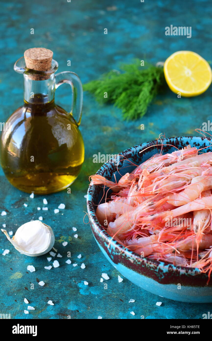 Raw shrimps in a traditional Greek bowl with olive oil jar, sea salt, lemon rosemary on a abstract background. Healthy eating concept Stock Photo