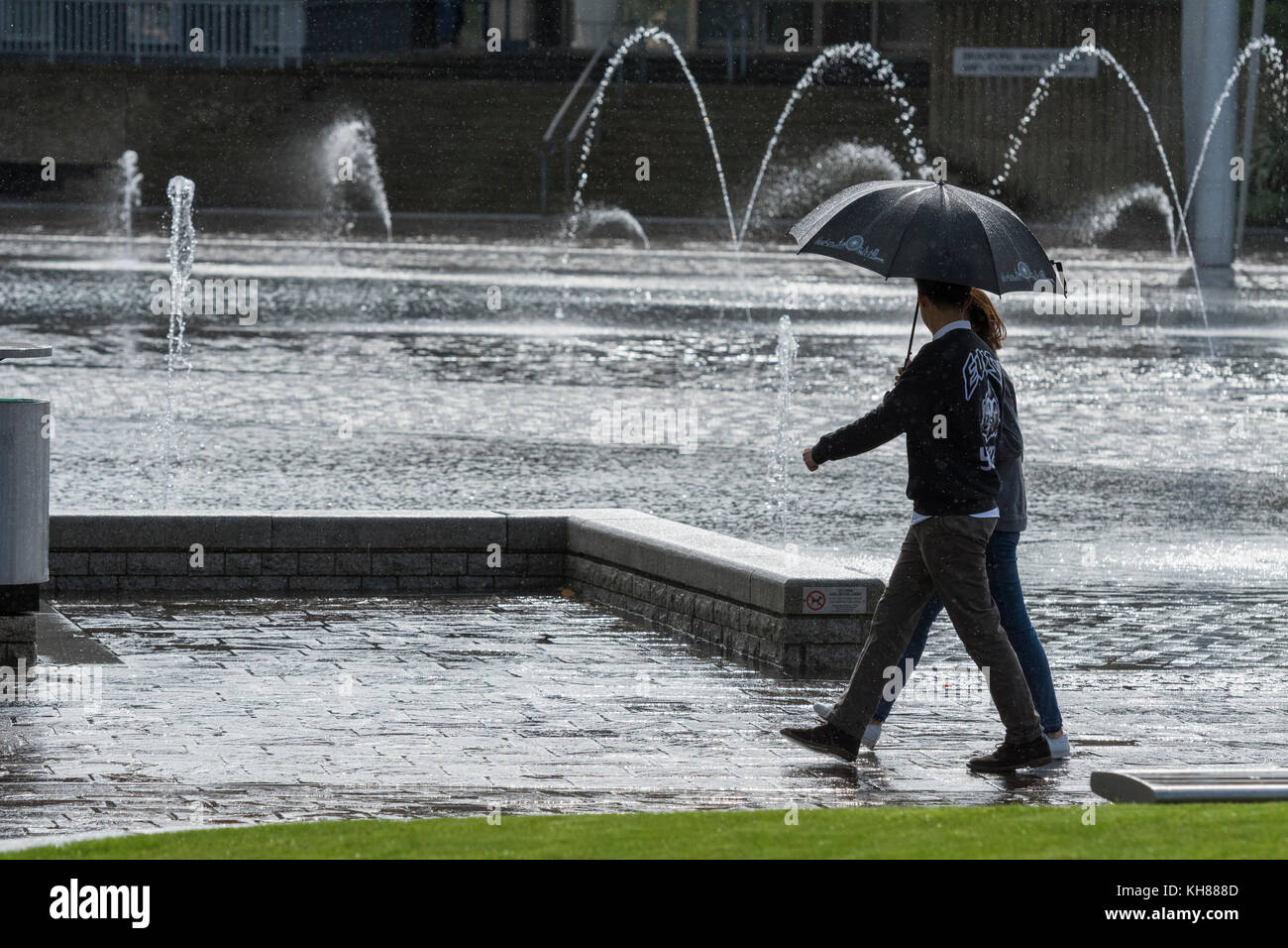 Couple walking in heavy rain, sheltering under umbrella, passing the Mirror Pool & fountains - Bradford centre City Park, West Yorkshire, England, UK. Stock Photo