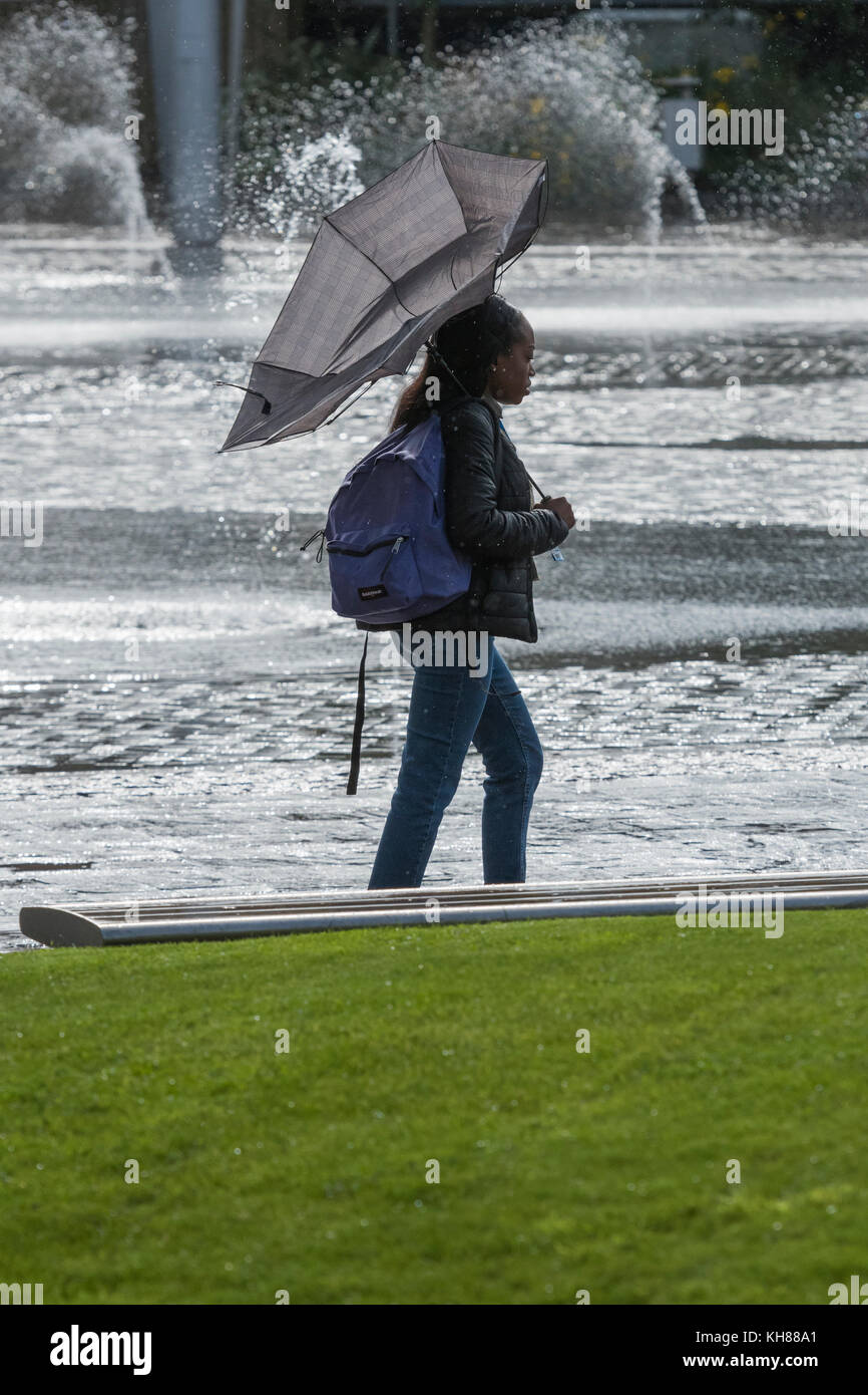 Woman walking in the heavy rain & wind, with brolly inside-out, passing the Mirror Pool & fountains - Bradford City Park, West Yorkshire, England, UK. Stock Photo