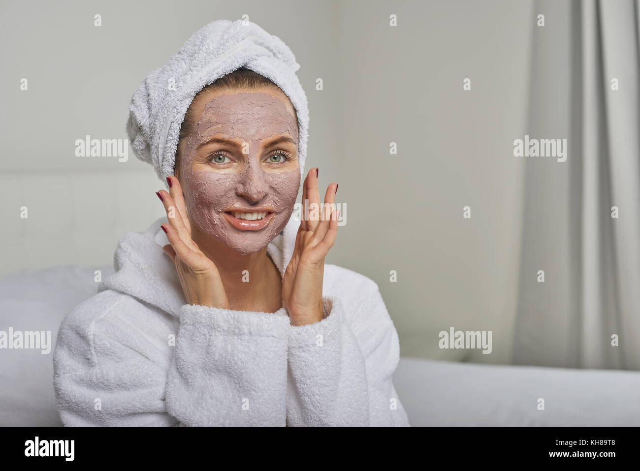 Woman at a spa having a face mask beauty treatment wearing white towelling robe and headdress smiling at the camera Stock Photo