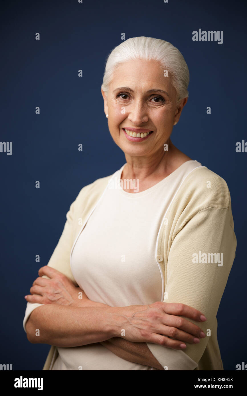 Photo of cheerful mature woman in beige tshirt, standing with crossed hands, looking at camera, over dark blue background Stock Photo