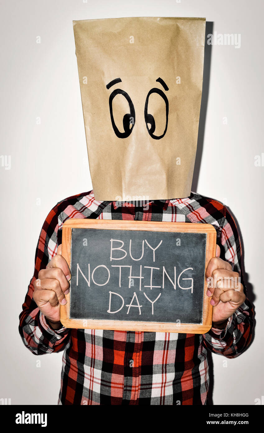 a young man wearing casual clothing with a paper bag in his head with a pair of eyes drawn in it shows a chalkboard with the text buy nothing day writ Stock Photo