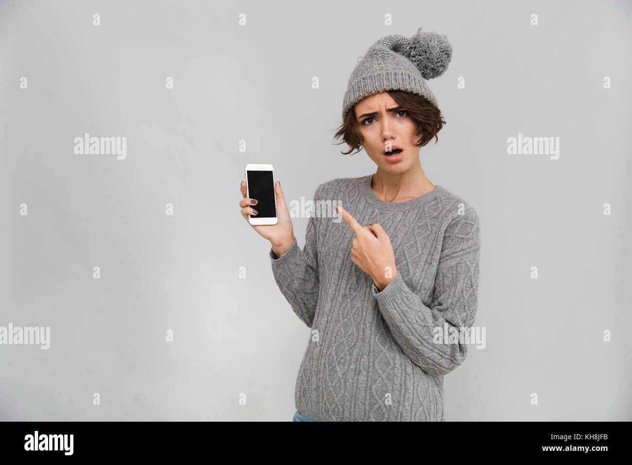 Porttrait of confused young girl in sweater and hat pointing finger at blank screen mobile phone isolated over gray background Stock Photo