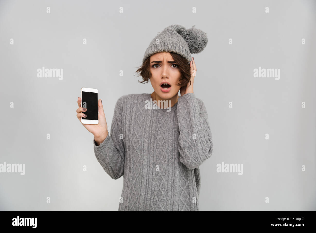Porttrait of shocked young girl in sweater and hat pointing finger at blank screen mobile phone isolated over gray background Stock Photo