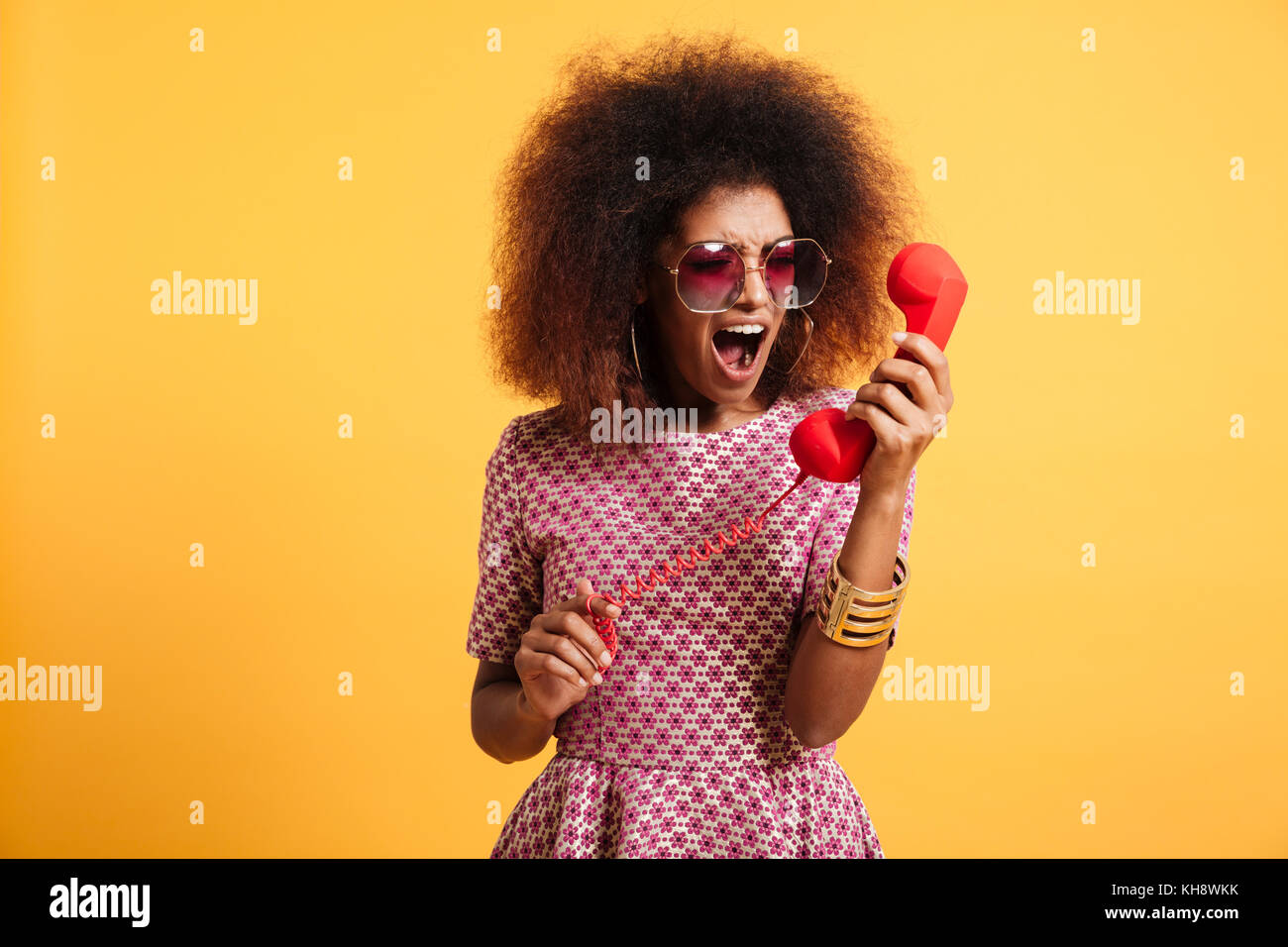 Portrait of a crazy angry afro american woman in retro style clothes yelling at telephone isolated over yellow background Stock Photo