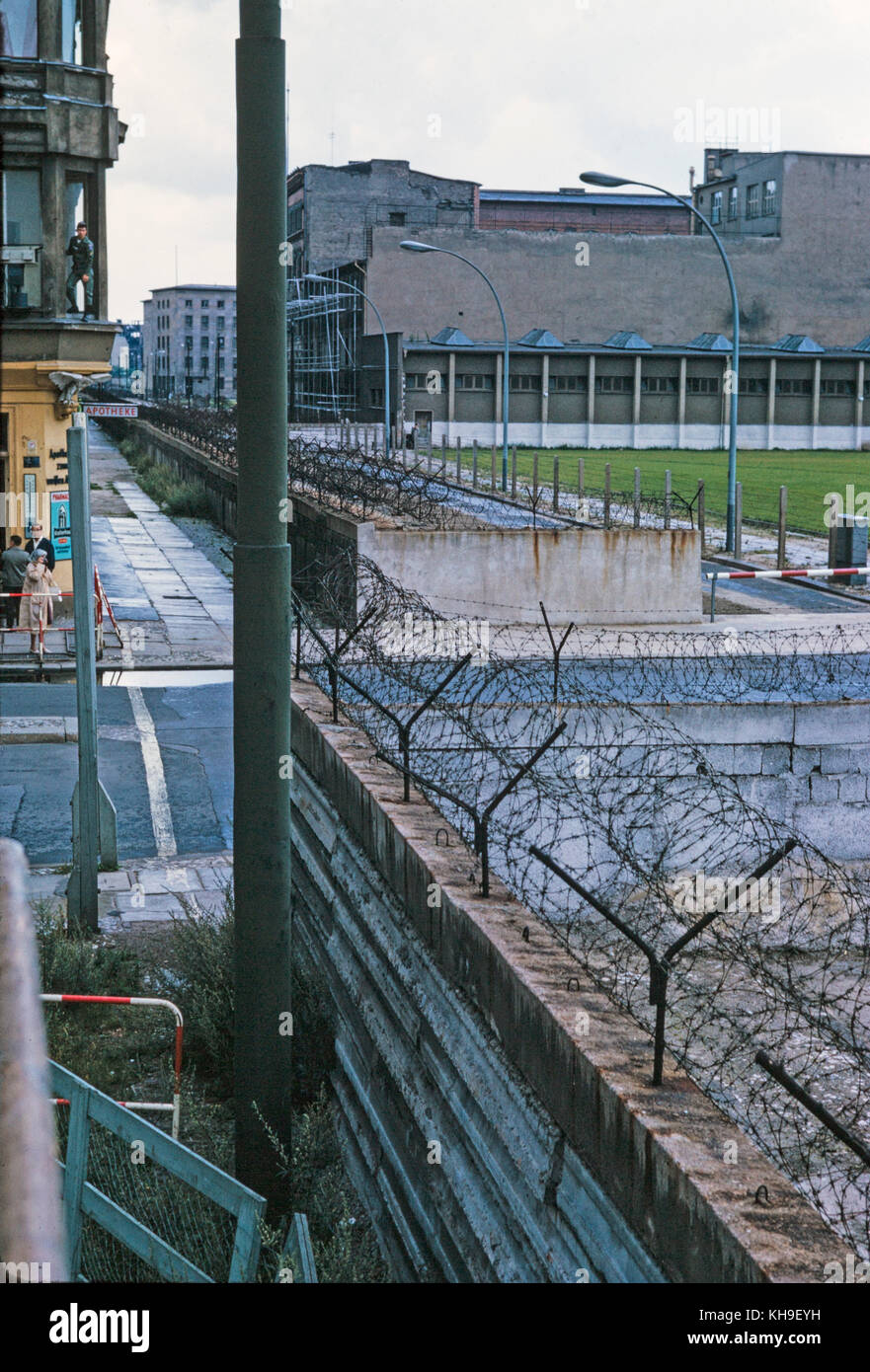 View looking across the Berlin Wall from West Germany in to East Germany in August 1965. The wall is covered in barbed wire and a female tourist is taking a photograph with a soldier appearing from an upstairs window of a nearby building. Stock Photo