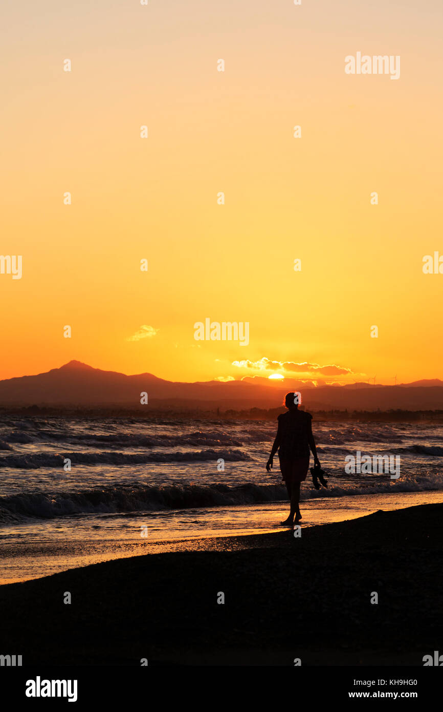 Woman walking in the surf, carrying shoes in the sunset. Stavrovouni hill in the background. Larnaca Cyprus Stock Photo