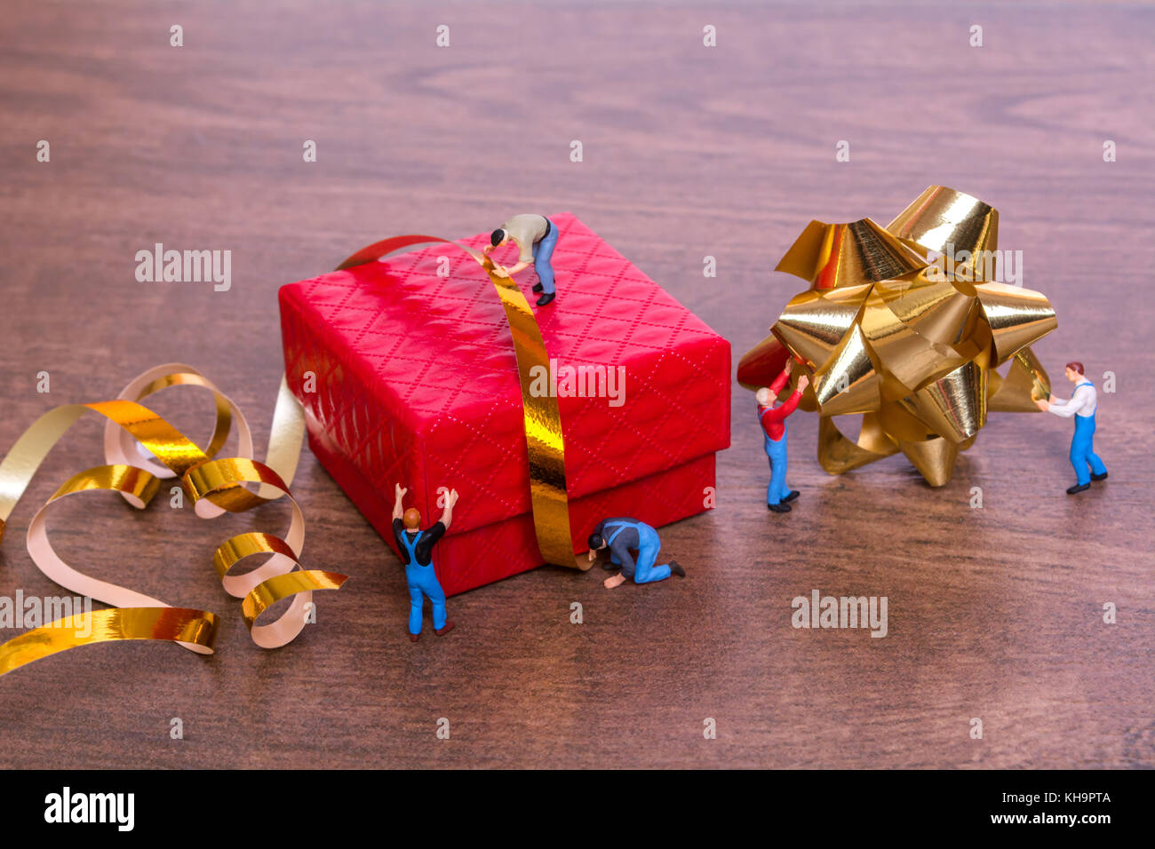 Creative concept with miniature people and a gift box on a wooden background. The process of packing gifts. Gold ribbon, bow, festive beads. Creative  Stock Photo