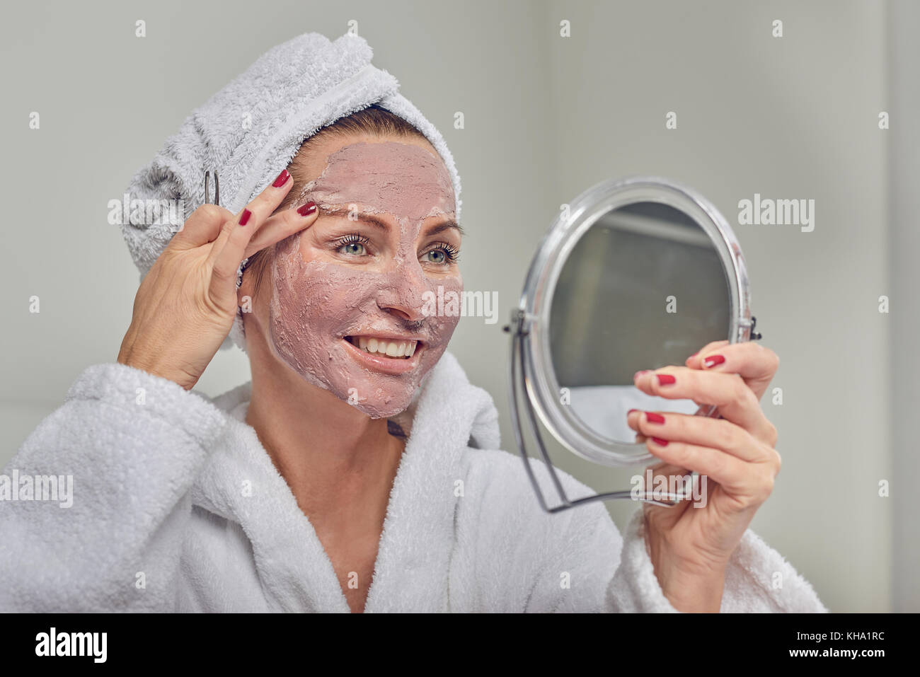 Attractive woman wearing a face mask beauty treatment holding a mirror as she plucks her eyebrows with tweezers Stock Photo