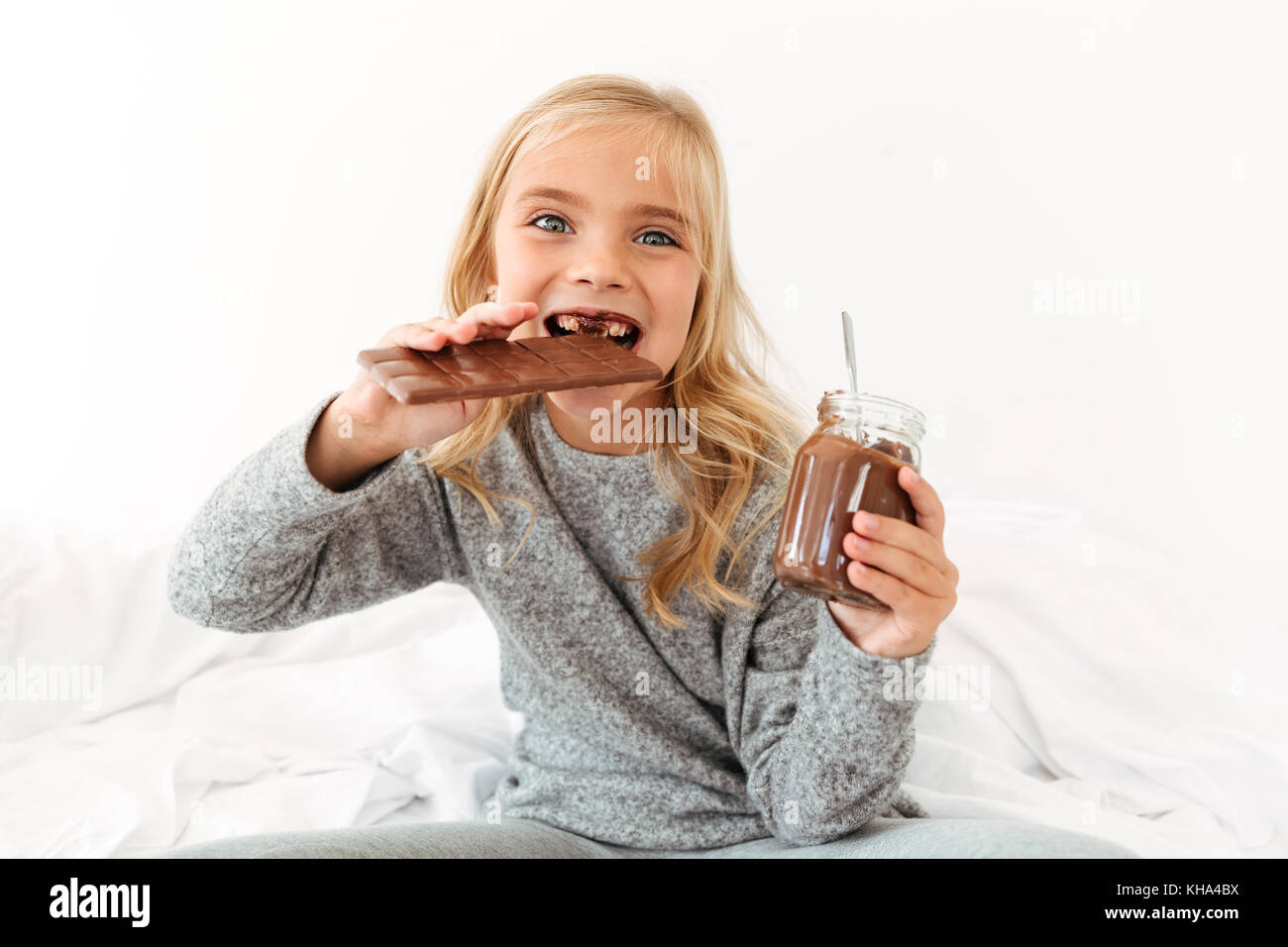 Close-up photo of funny little girl eating chocolate bar looking at camera while sitting on bed Stock Photo