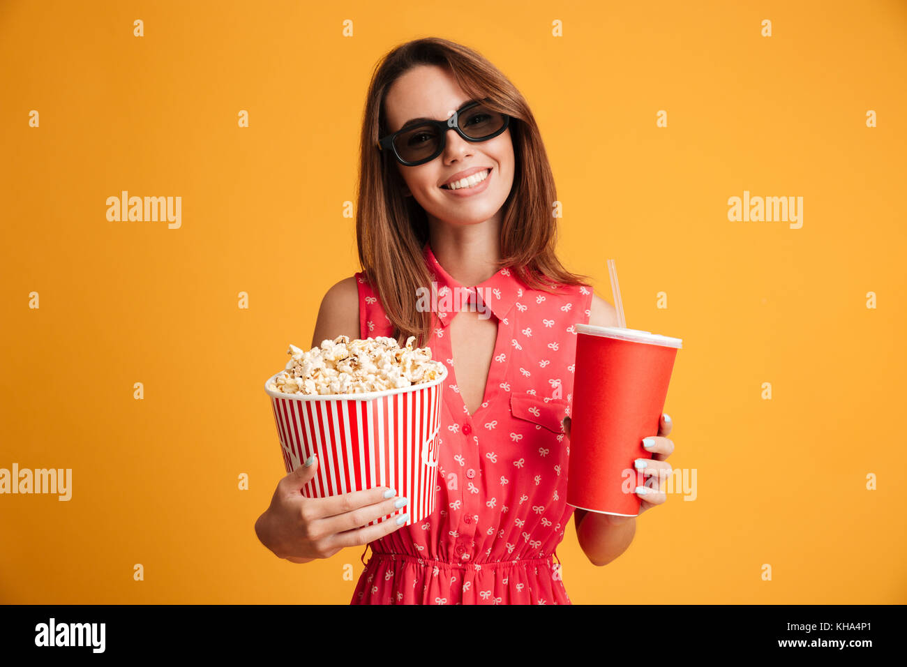Pleased brunette woman in dress and eyeglasses preparing to watch the film while holding popcorn and soda over yellow background Stock Photo