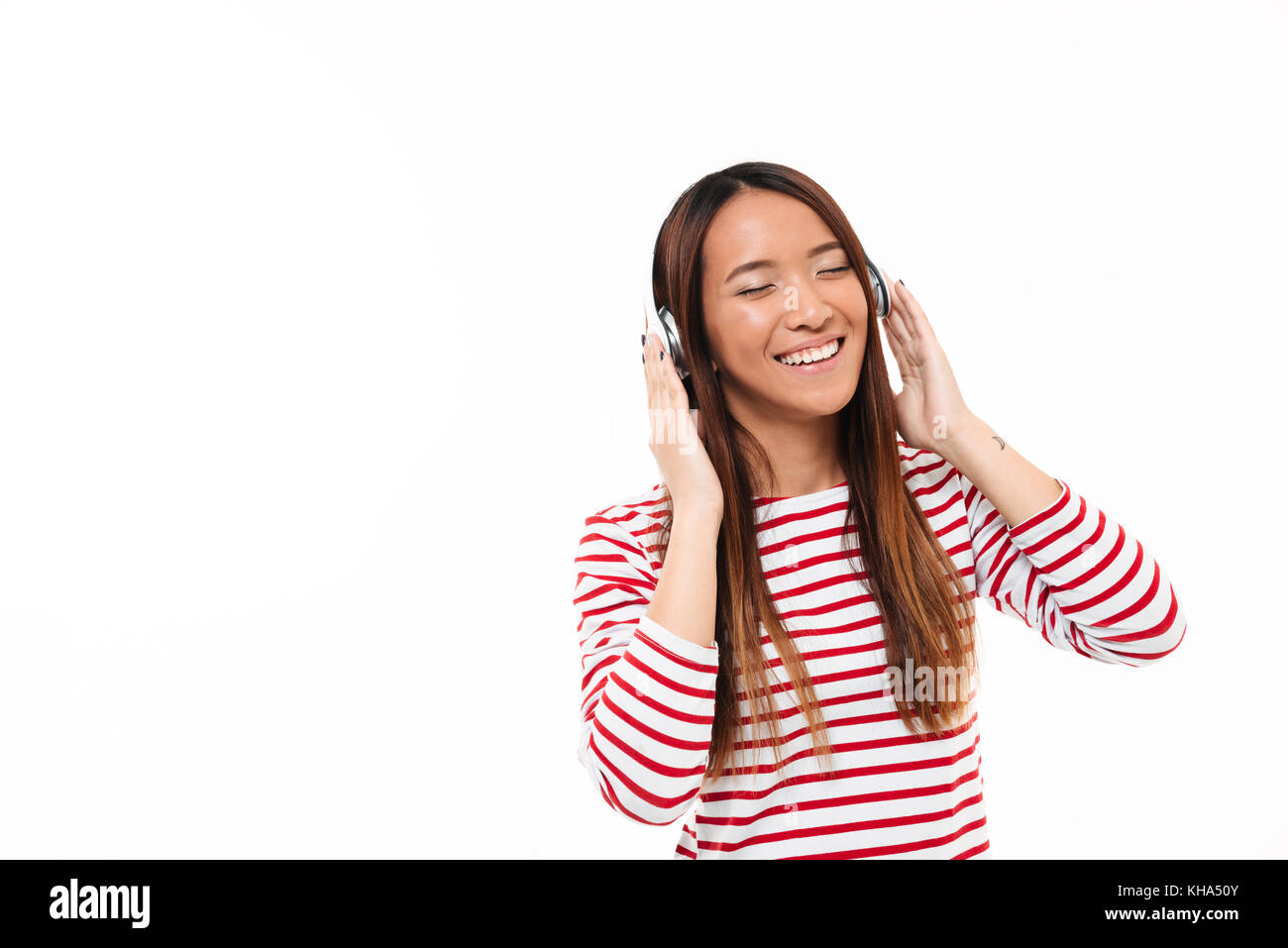 Portrait of a smiling joyful asian girl listening to music with headphones while standing isolated over white background Stock Photo