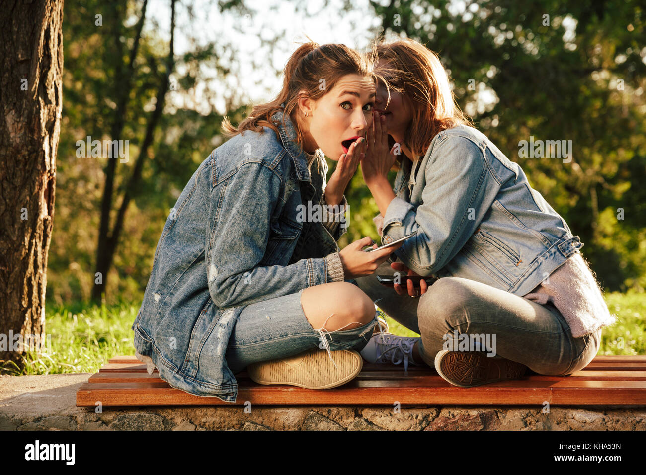 Two young brunette woman sharing gossips while sitting on wooden bench in  park Stock Photo - Alamy