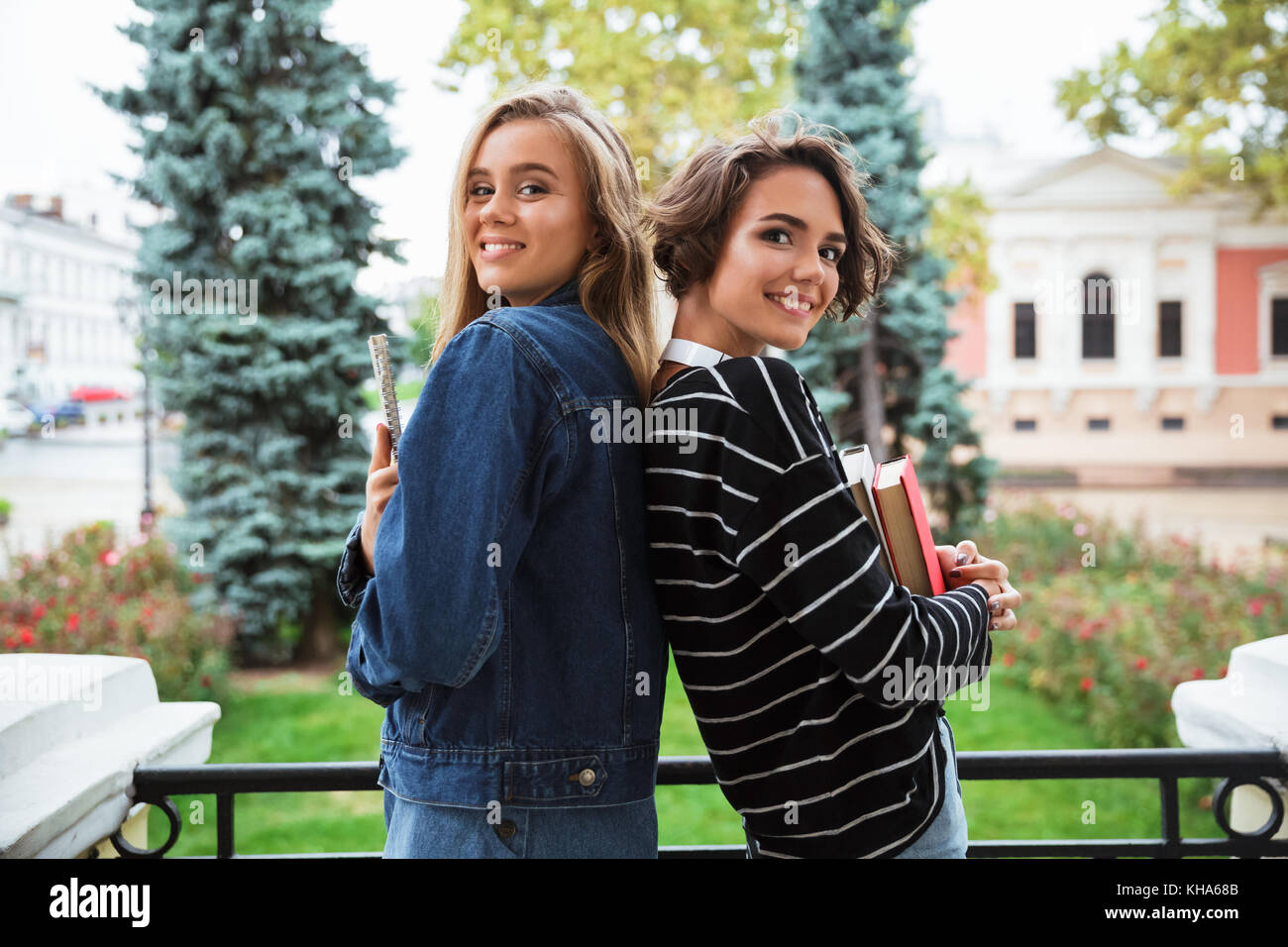 Two happy teenage girl holding books while standing back to back ...