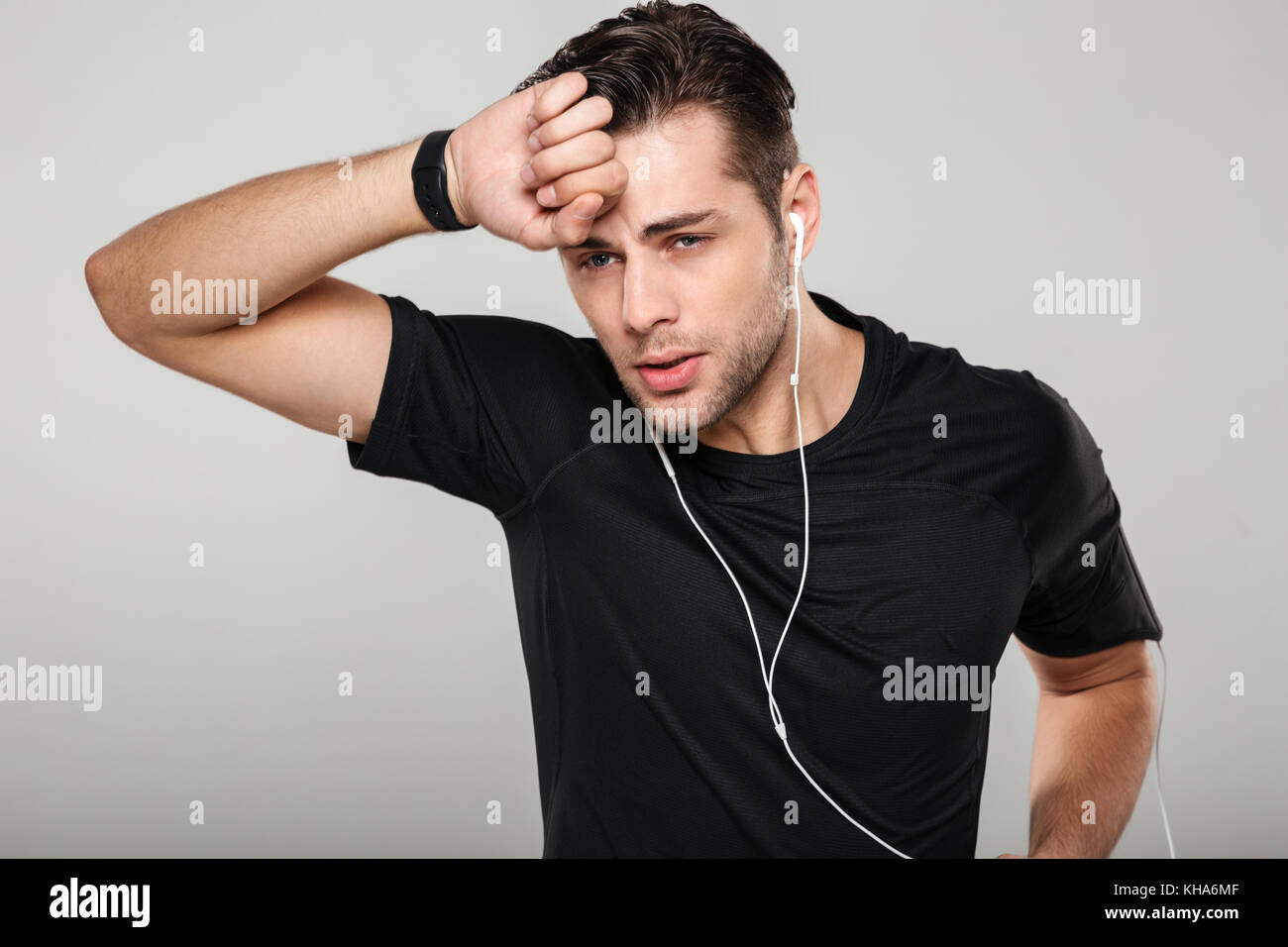 Portrait of a young tired sweaty sportsman in earphones wiping his forehead isolated over gray background Stock Photo