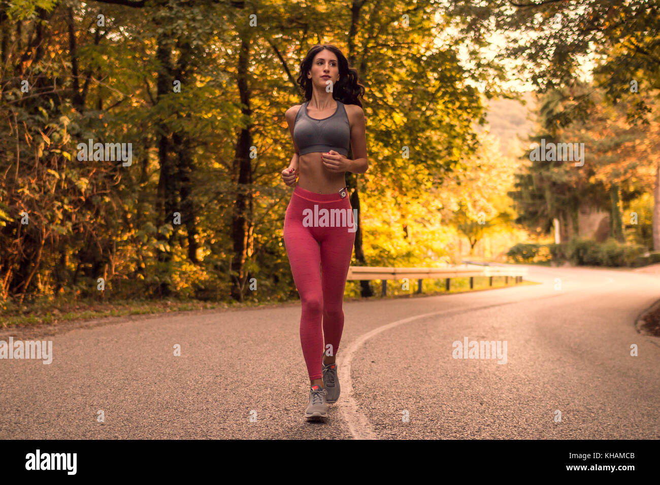 one young adult woman, running jogging, forest woods, asphalt road lane, sunset sunrise daylight, sport clothes Stock Photo