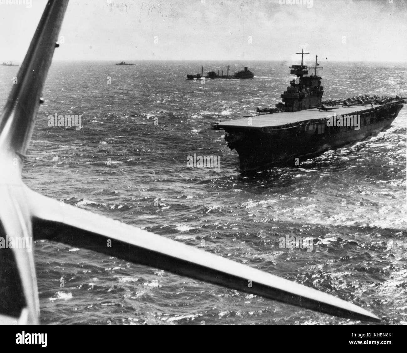 The U.S. Navy aircraft carrier USS Yorktown (CV-5) operating in the Pacific in February 1942, photographed from a Douglas TBD-1 torpedo plane that has just taken off from her deck. Other TBD and SBD aircraft are also ready to be launched. A F4F-3 'Wildcat' fighter is parked on the outrigger just forward of the island. The other ships in company include the fleet oiler USS Guadaloupe (AO-32), a destroyer and a heavy cruiser. Stock Photo
