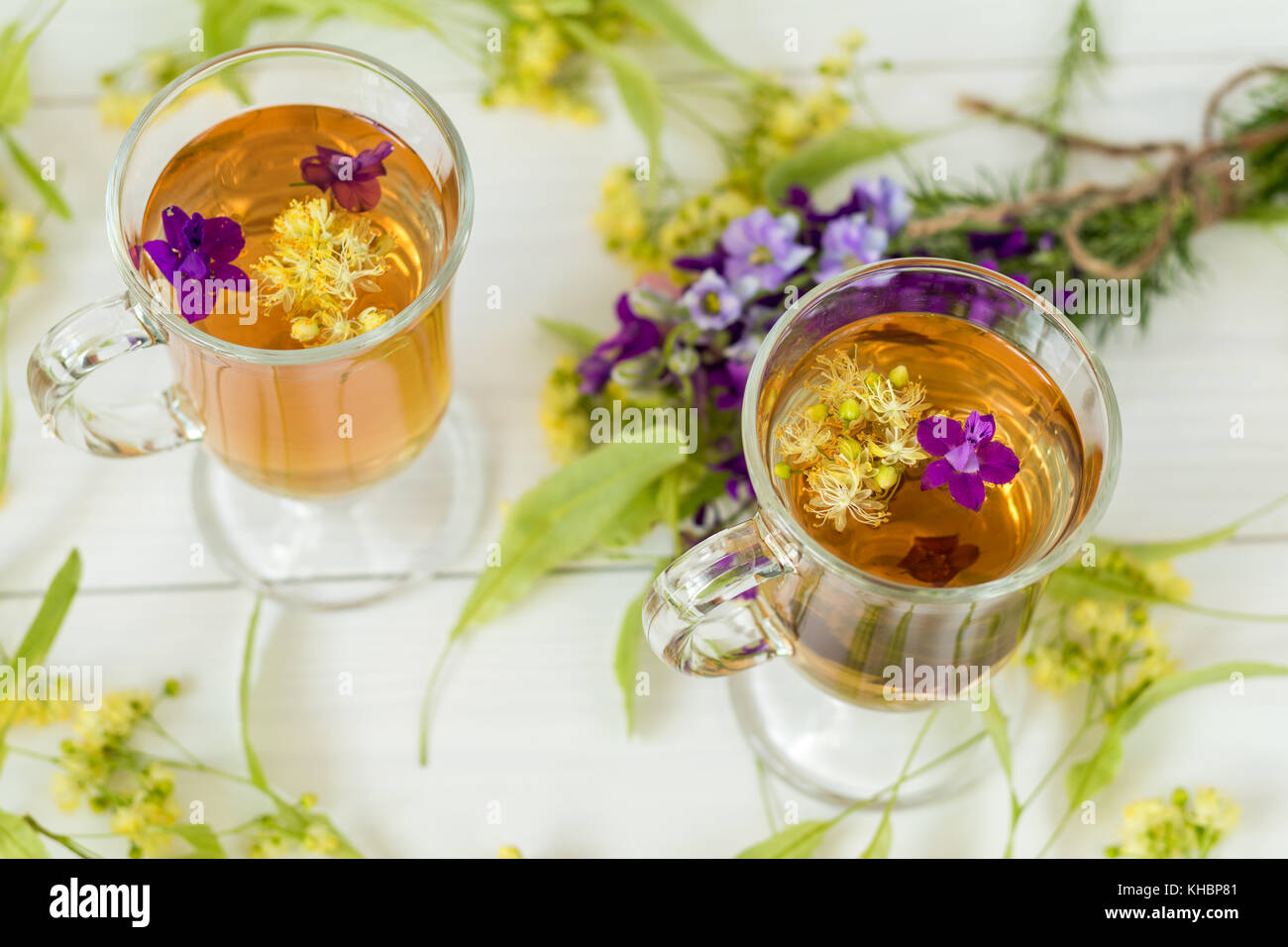 Linden herbal tea in a transparent grog glass with a linden blossom and bunch of herbs on the white wooden surface Stock Photo