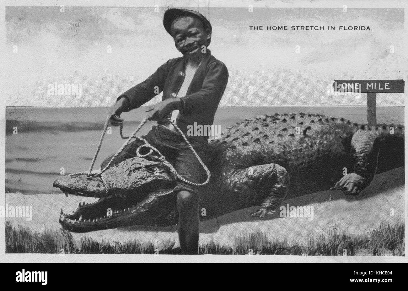 Hand colored postcard with an African American boy riding a crocodile on the sand, the ocean in the background, a sign that reads 'One Mile' behind them, titled 'The Home Stretch in Florida', 1914. From the New York Public Library. Stock Photo