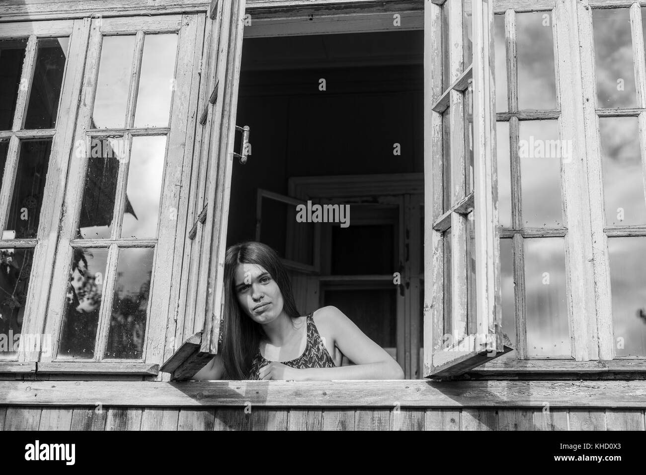 Young woman with long hair looks from of the window of the veranda. Black-and-white photo. Stock Photo