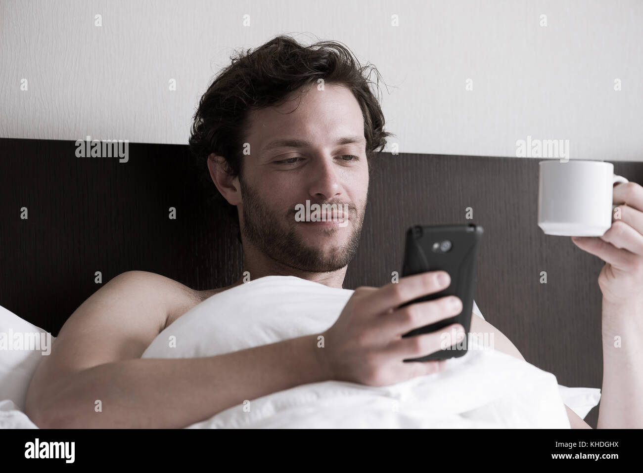 Man in bed using smartphone and drinking hot drink Stock Photo