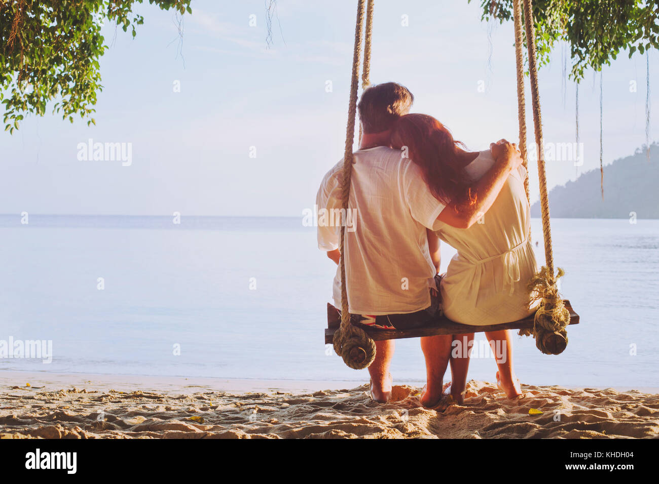 romantic holidays for two, affectionate couple sitting together on the beach on swing, silhouette of man hugging woman Stock Photo