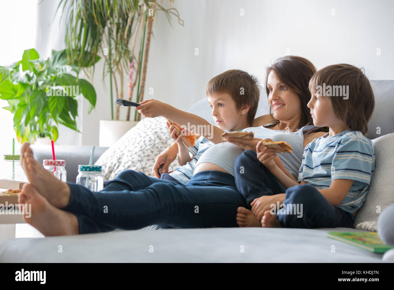 Happy young family, pregnant mother and two boys, eating tasty pizza at home, sitting on the sofa, watching TV and having a laugh Stock Photo