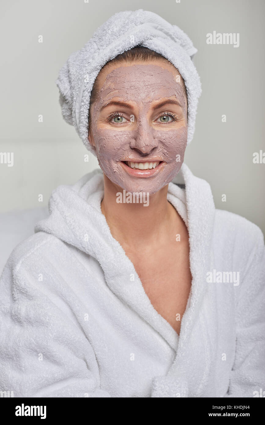 Woman at a spa having a face mask beauty treatment wearing white towelling robe and headdress smiling at the camera Stock Photo
